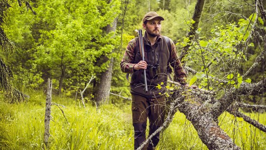 Mann im Wald, bekleidet mit Jagdausrüstung, steht auf einem umgefallenen Baumstamm und hält ein Gewehr in der Hand.
